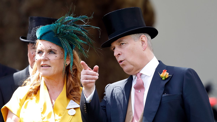 El príncipe Andrés con un traje azul oscuro y un sombrero de copa negro señala hacia arriba mientras Sarah Ferguson, con un vestido amarillo y un tocado verde, mira.