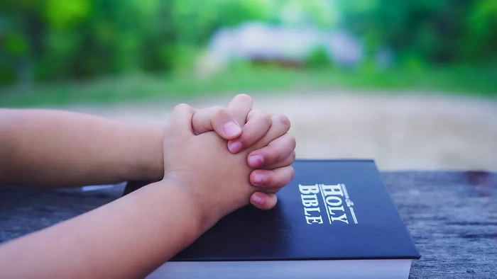 Las manos de un niño entrelazadas en oración descansan sobre una Biblia al aire libre.