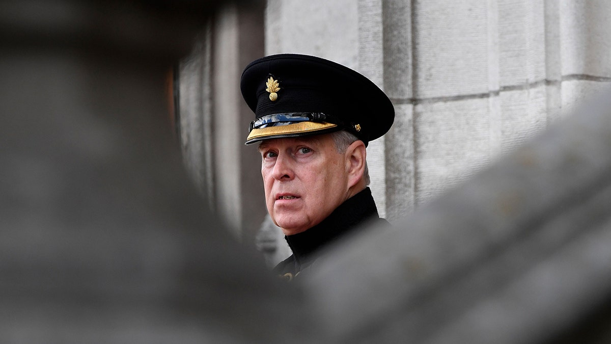 El príncipe Andrés con uniforme militar y sombrero mirando abatido detrás de un edificio gris.