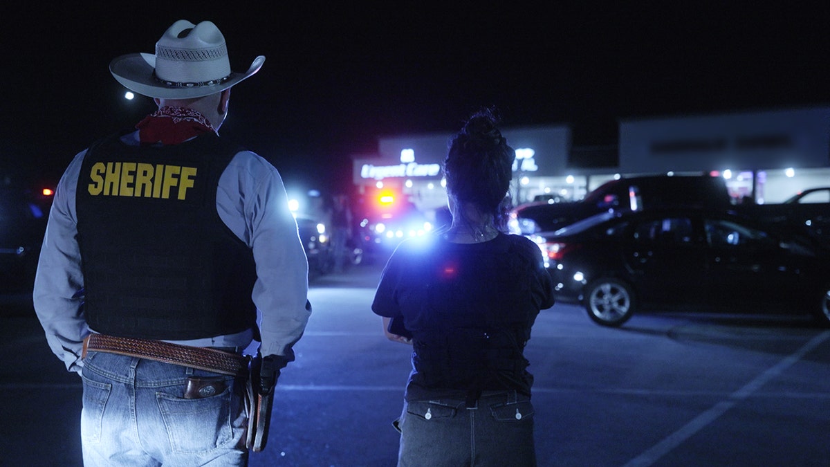 Roo Powell de pie junto a un sheriff por la noche
