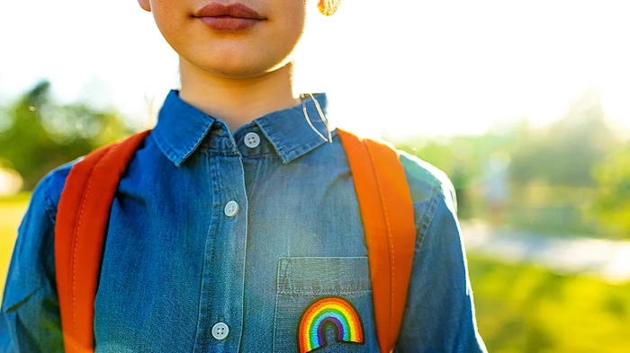 AAP сталкивается с давлением по поводу ухода за транс-молодежью, направляя врачей держать родителей в неведении относительно сексуального здоровья ребенка Девушка в джинсовой футболке с символом радуги и с рюкзаком стоит в летнем парке.