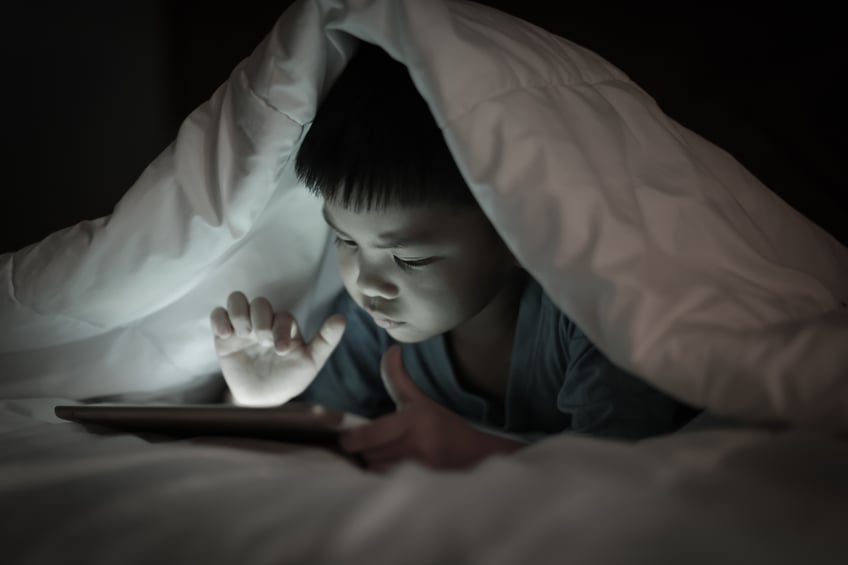 Picture of a child in bed covered by a sheet and using a tablet.