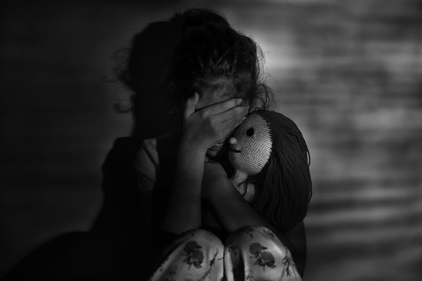 Girl hugging a doll, crying and hiding her face with her hands.