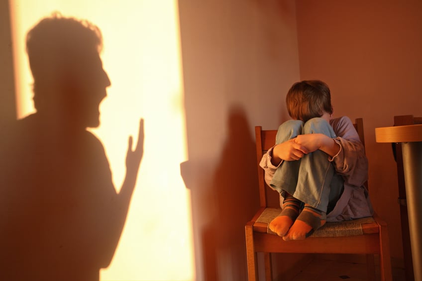 Shadow of a man shouting at a child sitting on a chair covering his face with his knees.